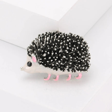 Hedgehog brooch with textured black and silver-tone spines, white body, pink ears and legs. Front view on white block in studio.