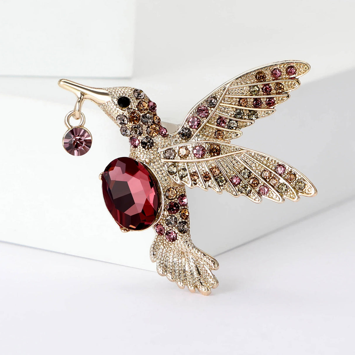 Hummingbird brooch with gold-tone metal, red faceted center stone, pink and brown decorative stones, angled view on white.