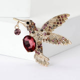 Hummingbird brooch with gold-tone metal, red faceted center stone, pink and brown decorative stones, angled view on white.