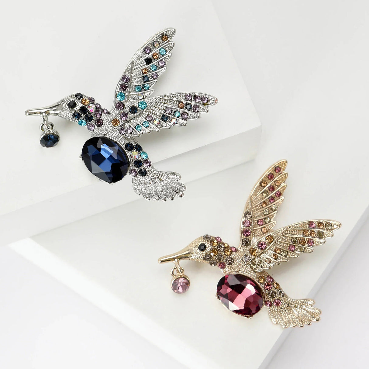 Pair of hummingbird brooches in silver-tone and gold-tone finishes, with blue and red faceted center stones and multicolor accents, flat lay on white.