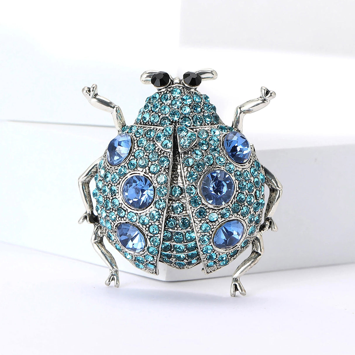Ladybug brooch with silver-tone metal, blue faceted stones, and black eyes, upright pose on white block.