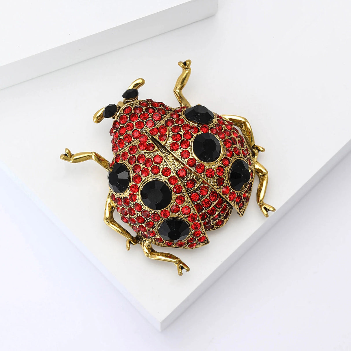 Ladybug brooch with gold-tone metal, red faceted stones, black decorative spots, and black eyes, angled view on white surface.