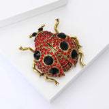 Ladybug brooch with gold-tone metal, red faceted stones, black decorative spots, and black eyes, angled view on white surface.