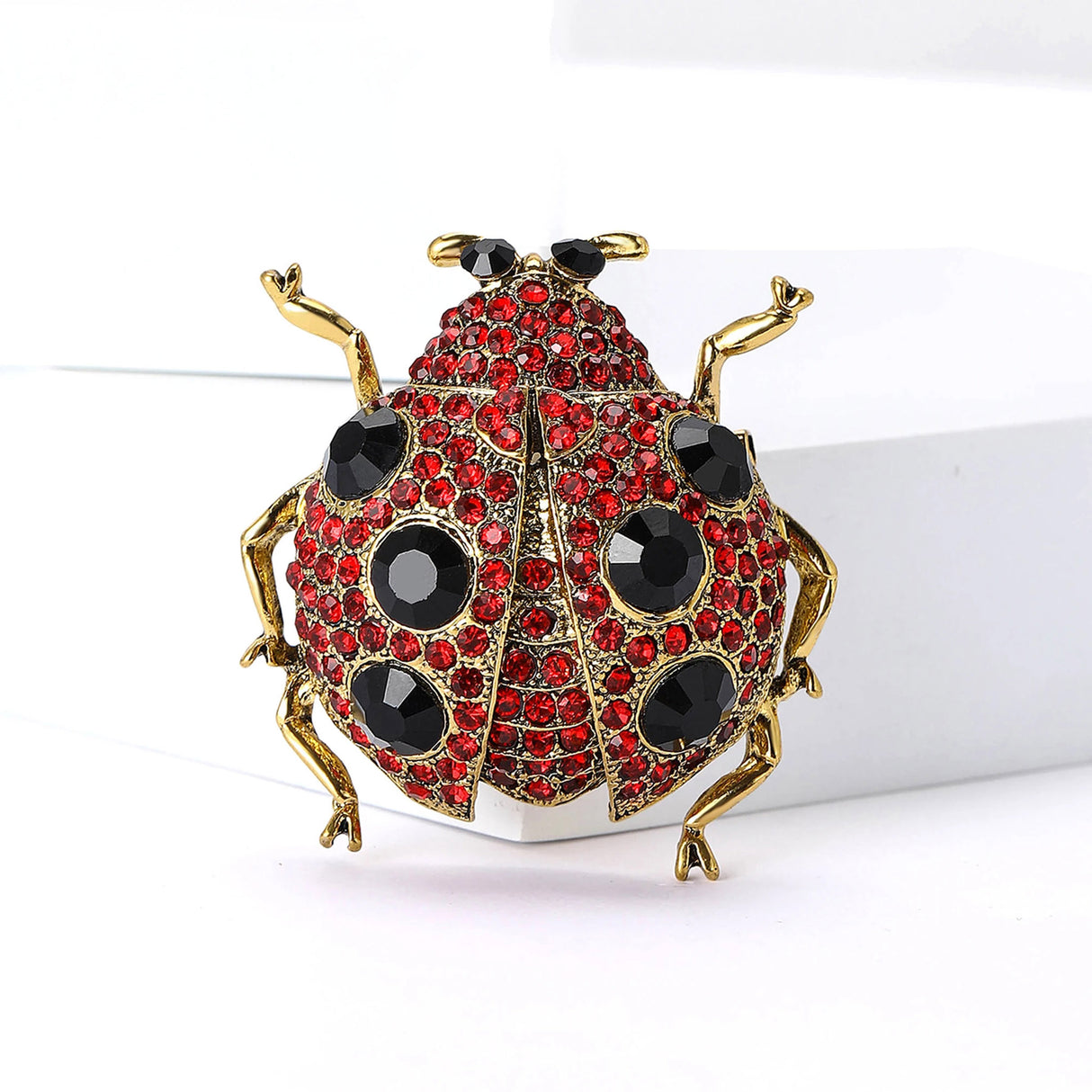 Ladybug brooch with gold-tone metal frame, red faceted stones, black decorative spots, and black eyes, upright front view on white block.