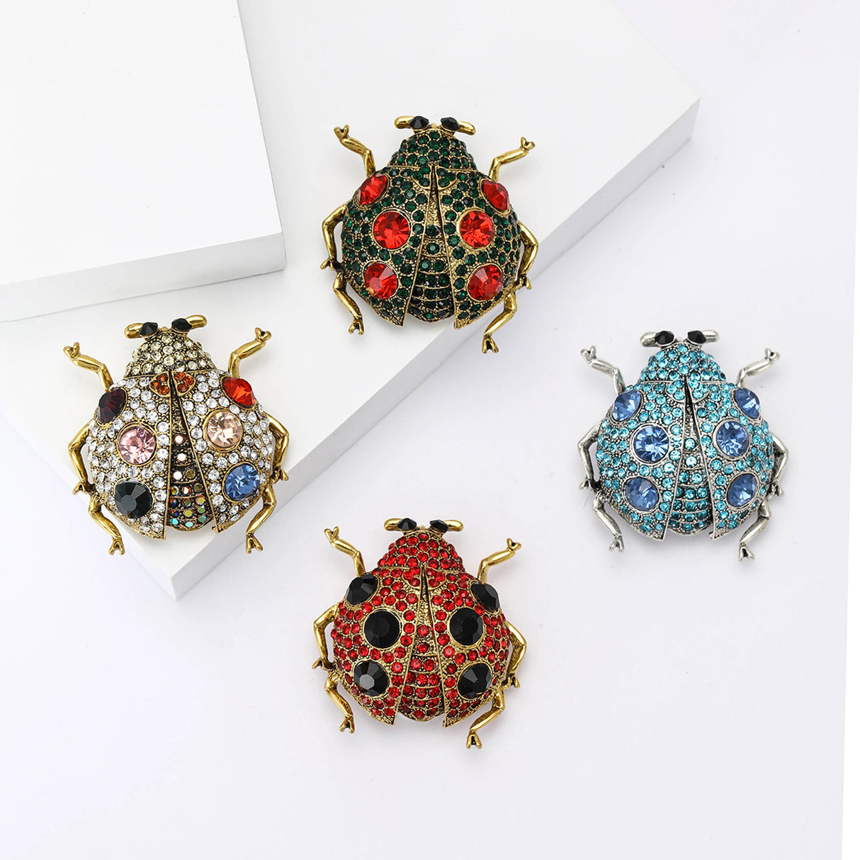 Set of four ladybird brooches in various colors, with faceted stones and metallic frames, flat lay on white blocks.