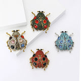Set of four ladybird brooches in various colors, with faceted stones and metallic frames, flat lay on white blocks.