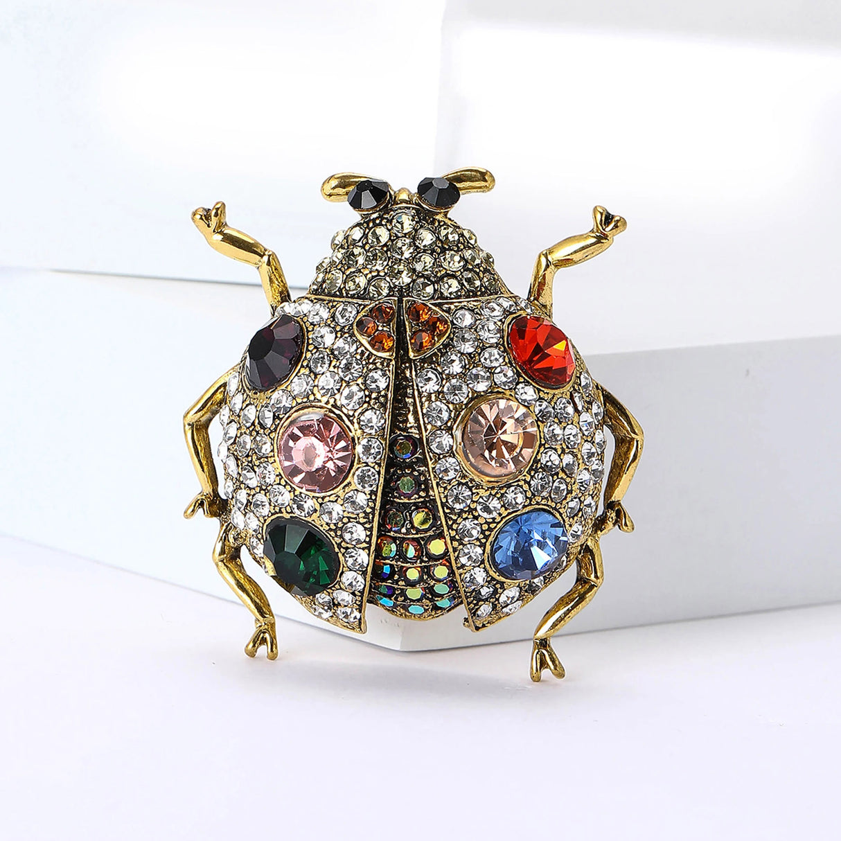 Ladybug brooch with gold-tone metal, clear base, multicolor faceted stones, and black eyes, upright front view on white block.