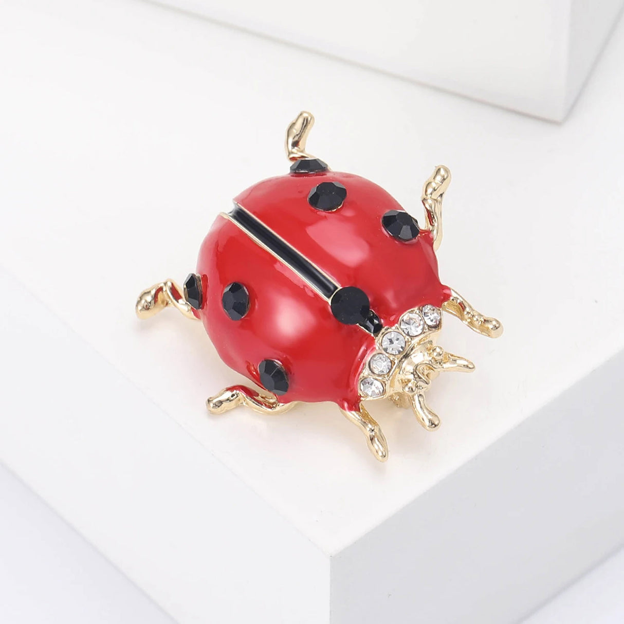 Ladybird brooch with red enamel body, black faceted spots, clear accent stones, and gold-tone legs, angled view on white block.