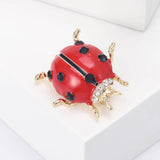 Ladybird brooch with red enamel body, black faceted spots, clear accent stones, and gold-tone legs, angled view on white block.