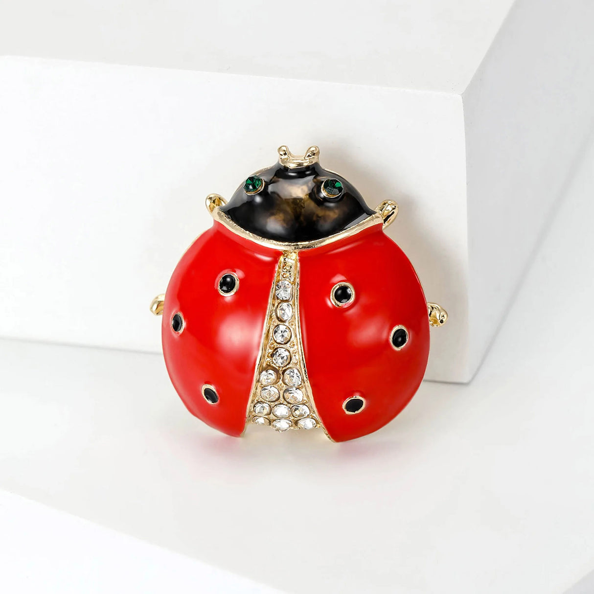 Ladybug brooch featuring red enamel wings, black circular spots, and clear decorative stones, angled view on white platform.