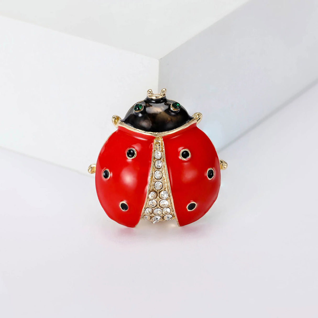 Ladybird brooch with red enamel finish, gold-tone accents, black head, and clear stones, upright view on white block.