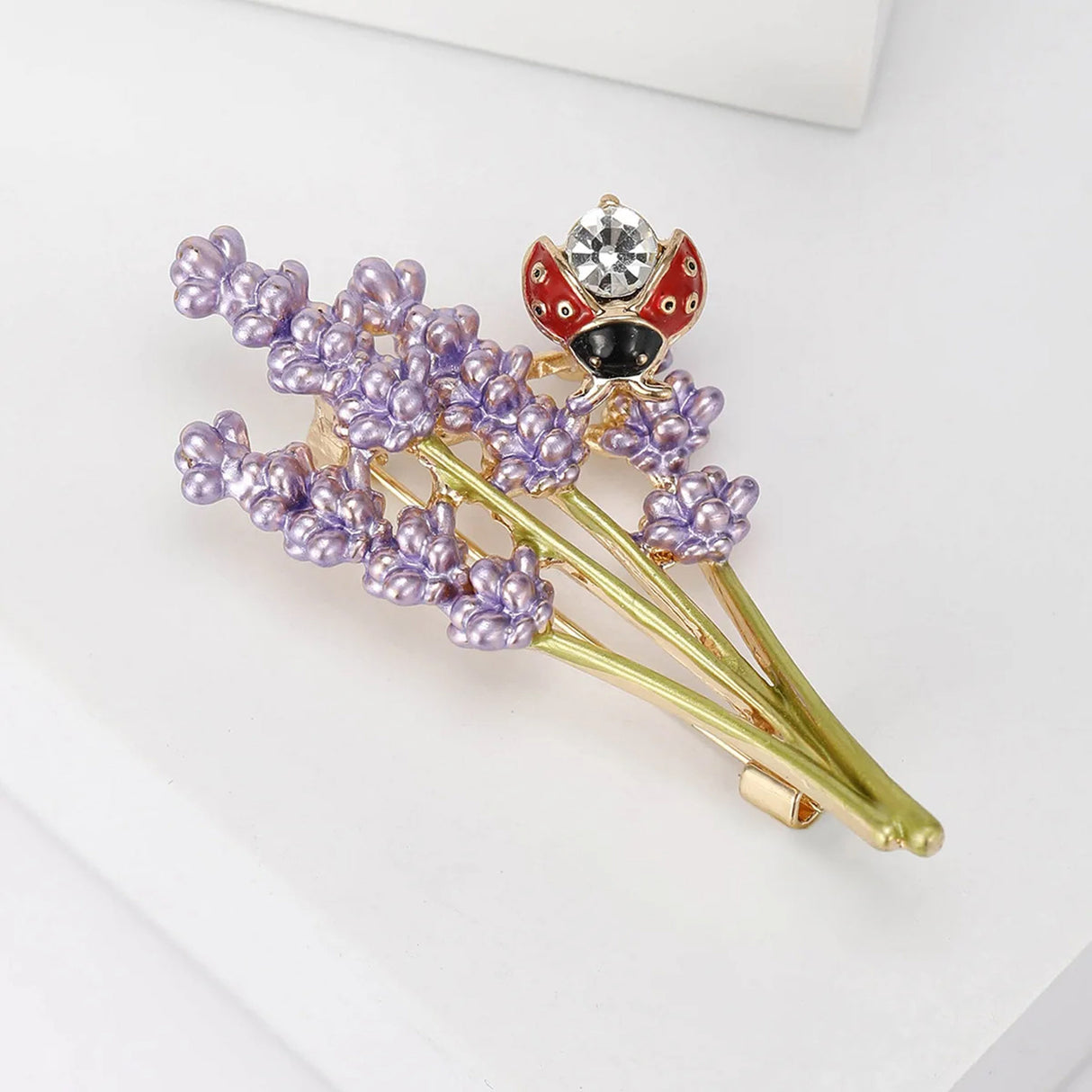 Lavender flower brooch featuring purple enamel buds, gold-tone stems, and red ladybird with clear stone, flat lay on white studio backdrop.