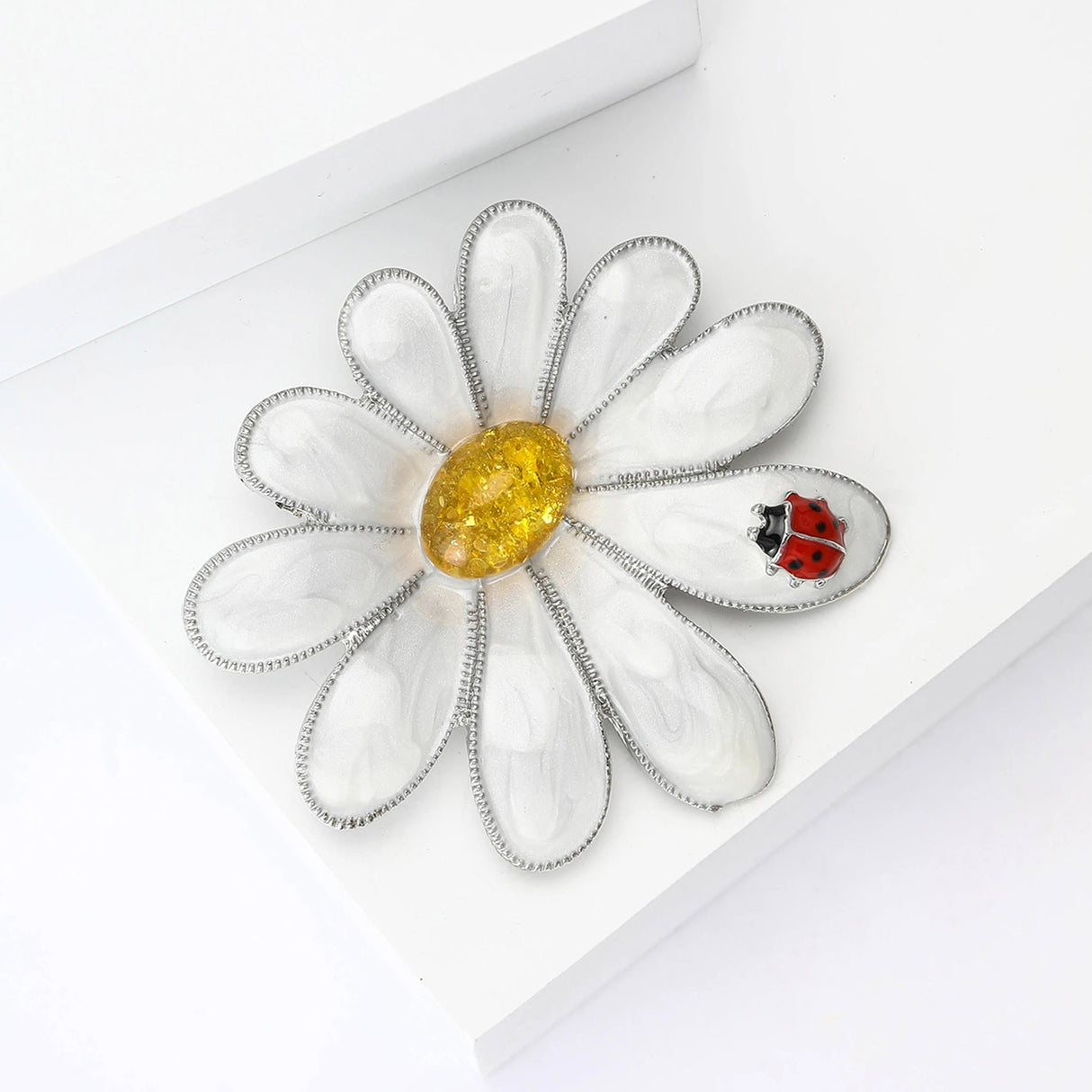 Daisy brooch with silver-tone edging, white enamel petals, yellow stone center, and red ladybird motif, angled flat lay.