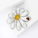 Daisy brooch with silver-tone edging, white enamel petals, yellow stone center, and red ladybird motif, angled flat lay.