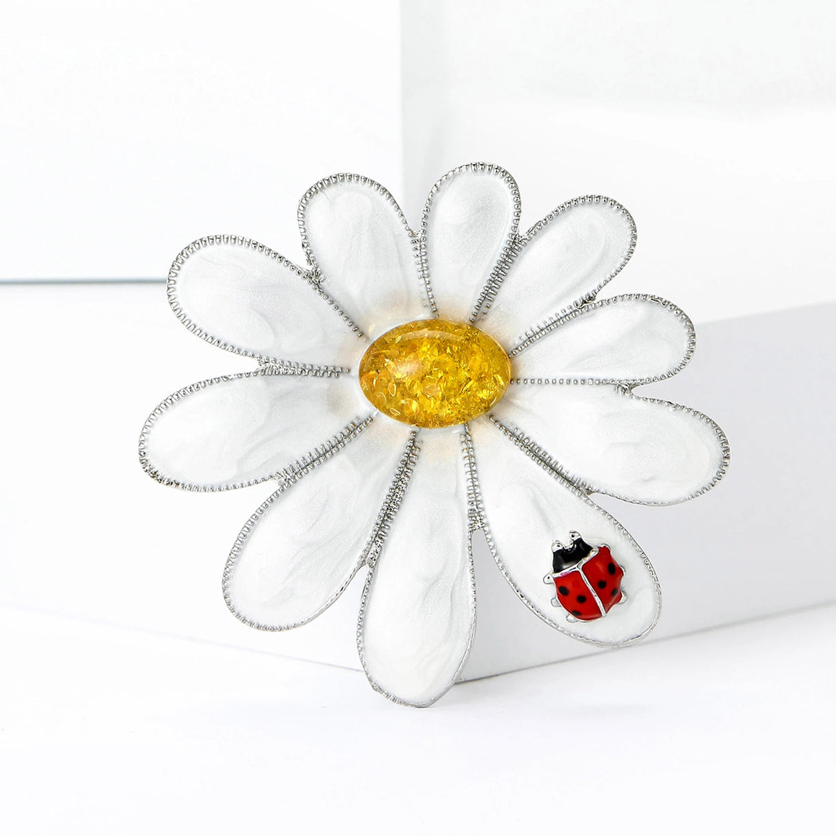 Daisy brooch with silver-tone border, glossy white petals, yellow stone center, and red ladybug accent, upright on white block.