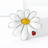 Daisy brooch with silver-tone border, glossy white petals, yellow stone center, and red ladybug accent, upright on white block.