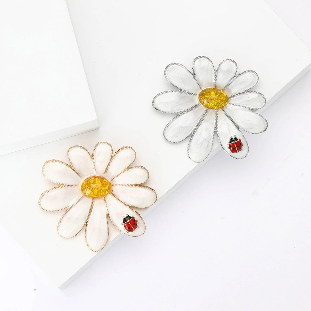 Pair of daisy brooches in gold-tone and silver-tone metal finishes with white enamel petals, yellow stone centers, and red ladybird accents. White studio flat lay.