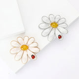 Pair of daisy brooches in gold-tone and silver-tone metal finishes with white enamel petals, yellow stone centers, and red ladybird accents. White studio flat lay.