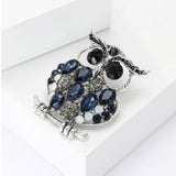 Owl brooch with silver-tone finish, dark blue teardrop stones, clear round accents, and black eyes, angled view on white blocks.