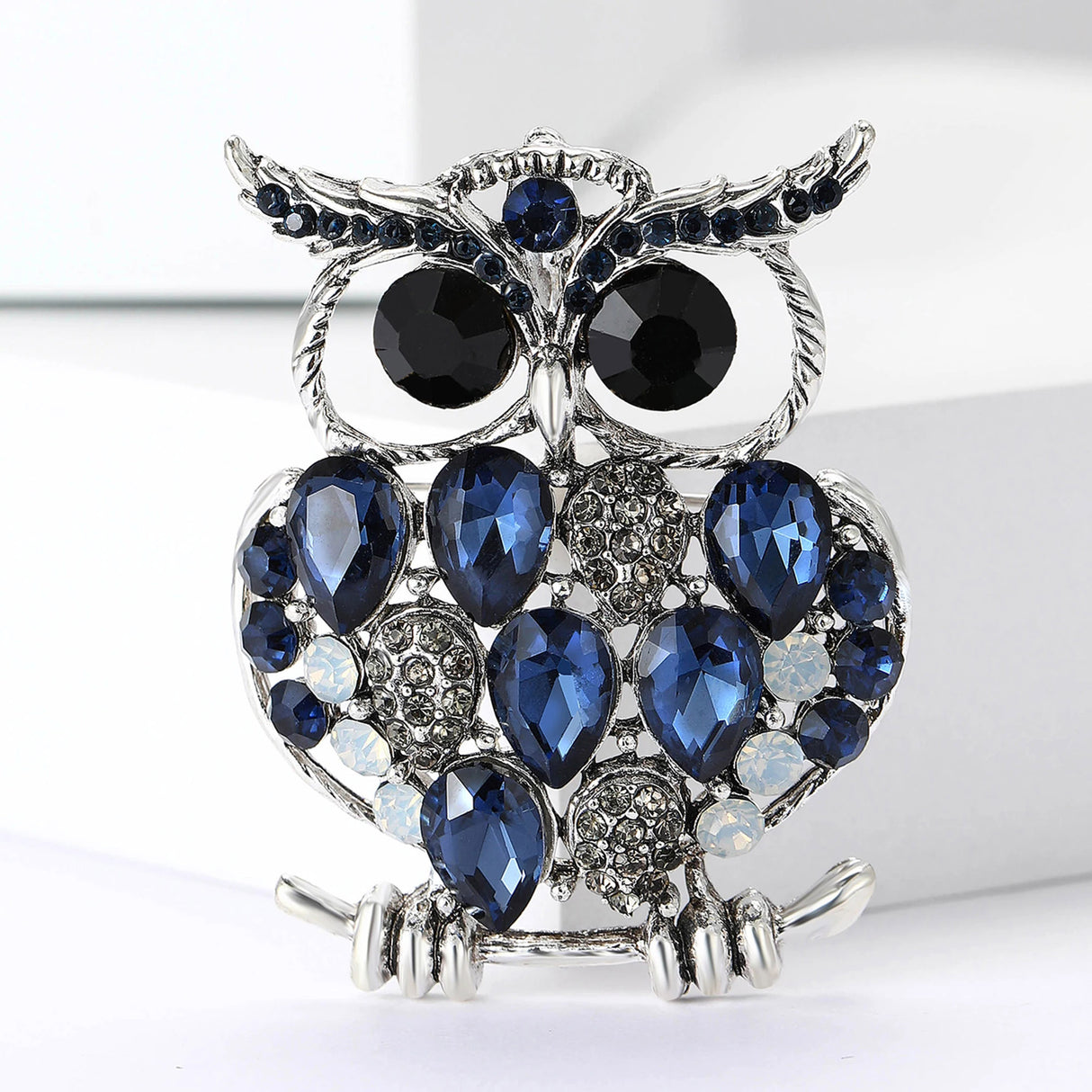 Owl brooch with silver-tone metal, blue faceted stones, black round eyes, and white accent stones, front view on white surface.