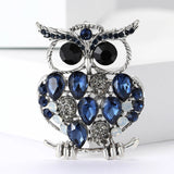 Owl brooch with silver-tone metal, blue faceted stones, black round eyes, and white accent stones, front view on white surface.