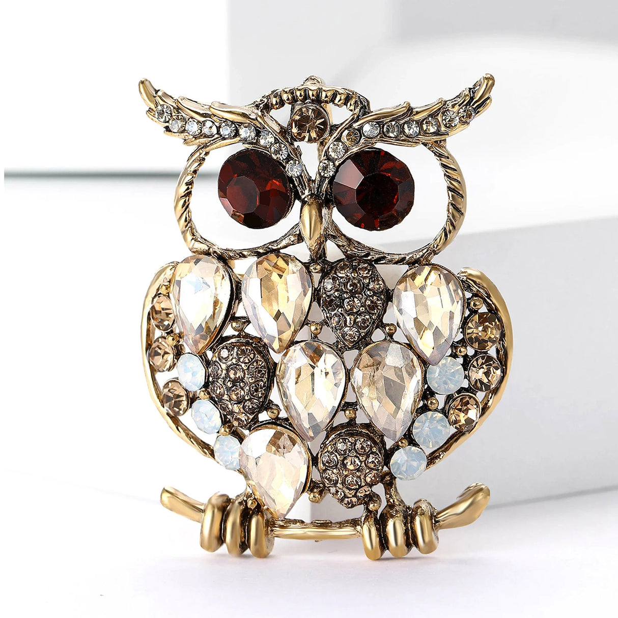 Owl brooch with gold-tone finish, beige teardrop stones, red eyes, and round white accents, front view on white background.