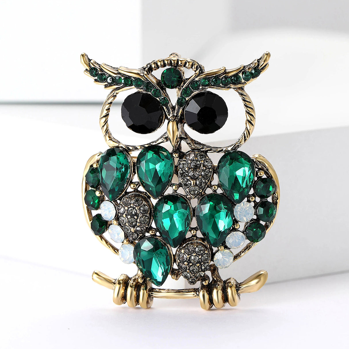 Owl brooch with gold-tone base, green faceted teardrop stones, black round eyes, and white round accents, front view on white blocks.