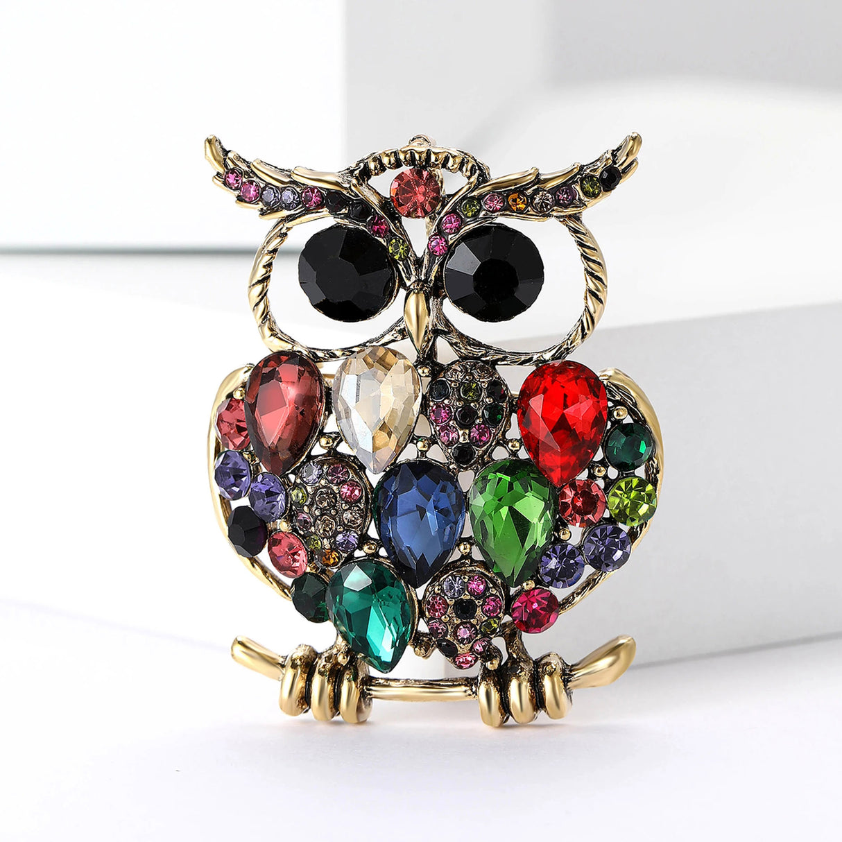 Owl brooch with gold-tone finish, multicolor faceted stones including red, green, blue, and pink tones; black eyes. Front view on white surface.