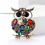 Owl brooch with gold-tone finish, multicolor faceted stones including red, green, blue, and pink tones; black eyes. Front view on white surface.