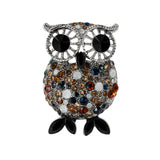 Owl brooch with silver-tone frame, large black eyes, and multicolor faceted stones including brown, amber, blue and white, with black oval stones with pointed tips for feet, cutout on white.