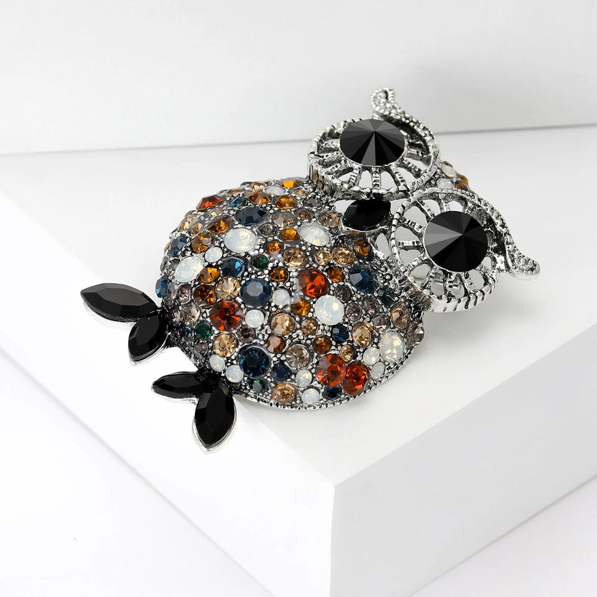 Owl brooch with silver-tone frame, black eyes, and round faceted stones in amber, blue, white, and brown, with black leaf-shaped feet, side view on white 