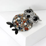 Owl brooch with silver-tone frame, black eyes, and round faceted stones in amber, blue, white, and brown, with black leaf-shaped feet, side view on white 