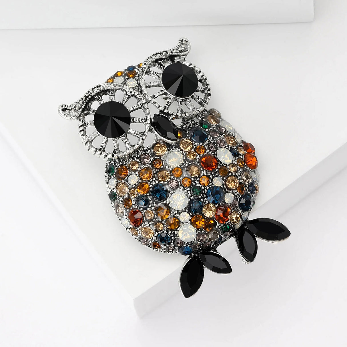 Owl brooch featuring silver-tone detailing, black eyes, and multicolor round stones including blue, orange, beige, and white, with black elongated oval feet, resting at an angle on white block.