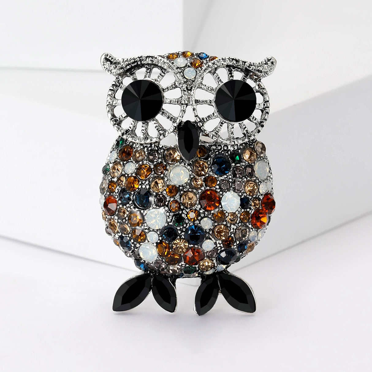 Owl brooch with silver-tone metal, large black eyes, and round pave stones in shades of brown, blue, and white, finished with black pointed oval feet, upright view on white background.