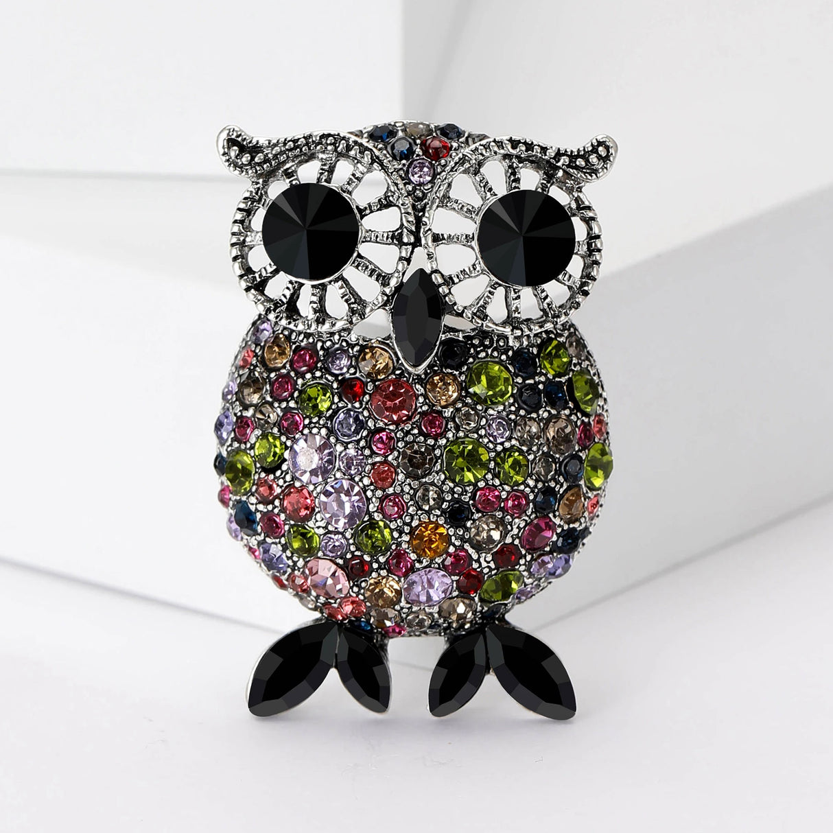 Owl brooch with silver-tone frame, black eyes, and round faceted stones including pink, green, red, and purple colors. Black tapered oval stones for feet. Upright view on layered white platform.