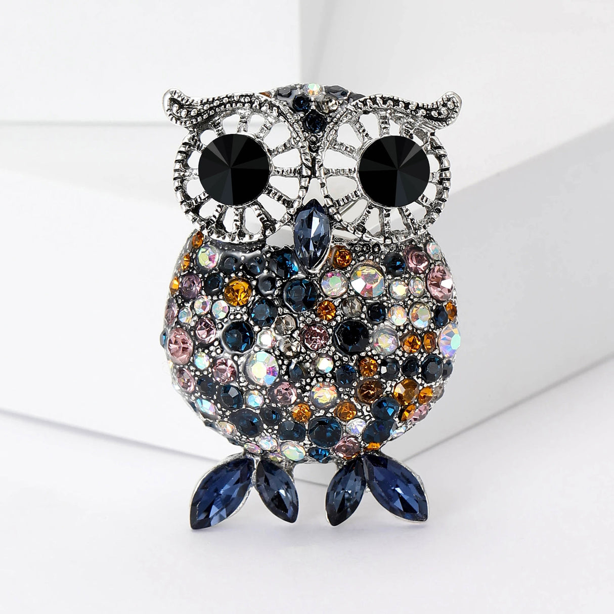 Owl brooch with silver-tone setting, large black eyes, and faceted stones in blue, orange, pink, and iridescent tones, with dark blue navette-shaped feet, standing on white surface.