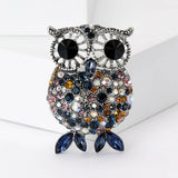 Owl brooch with silver-tone setting, large black eyes, and faceted stones in blue, orange, pink, and iridescent tones, with dark blue navette-shaped feet, standing on white surface.