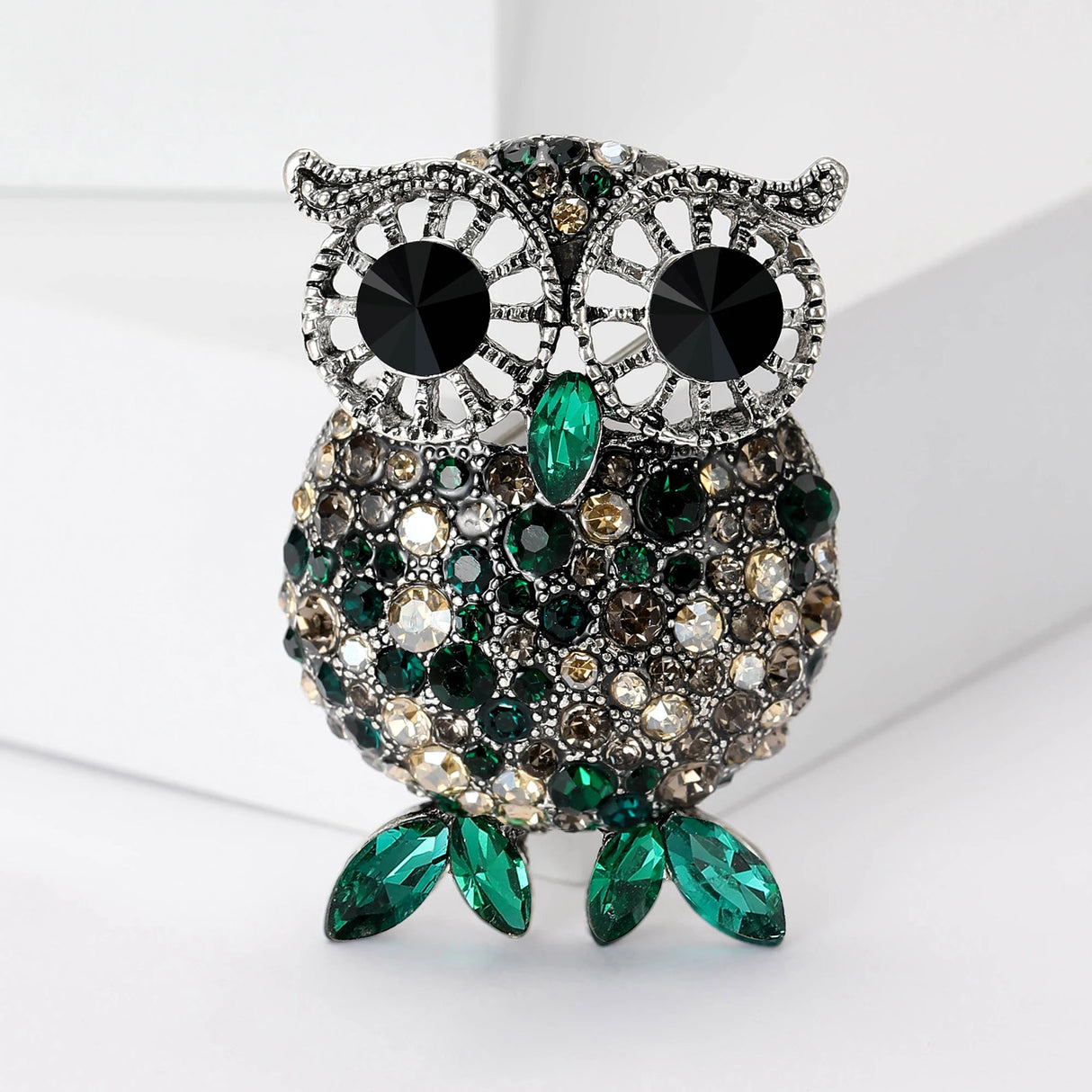 Owl brooch featuring silver-tone frame, black eyes, and faceted stones in green, beige, and gray tones, with green leaf-like stones for feet, upright on white angled block.