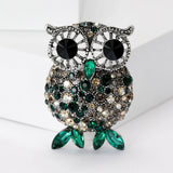 Owl brooch featuring silver-tone frame, black eyes, and faceted stones in green, beige, and gray tones, with green leaf-like stones for feet, upright on white angled block.