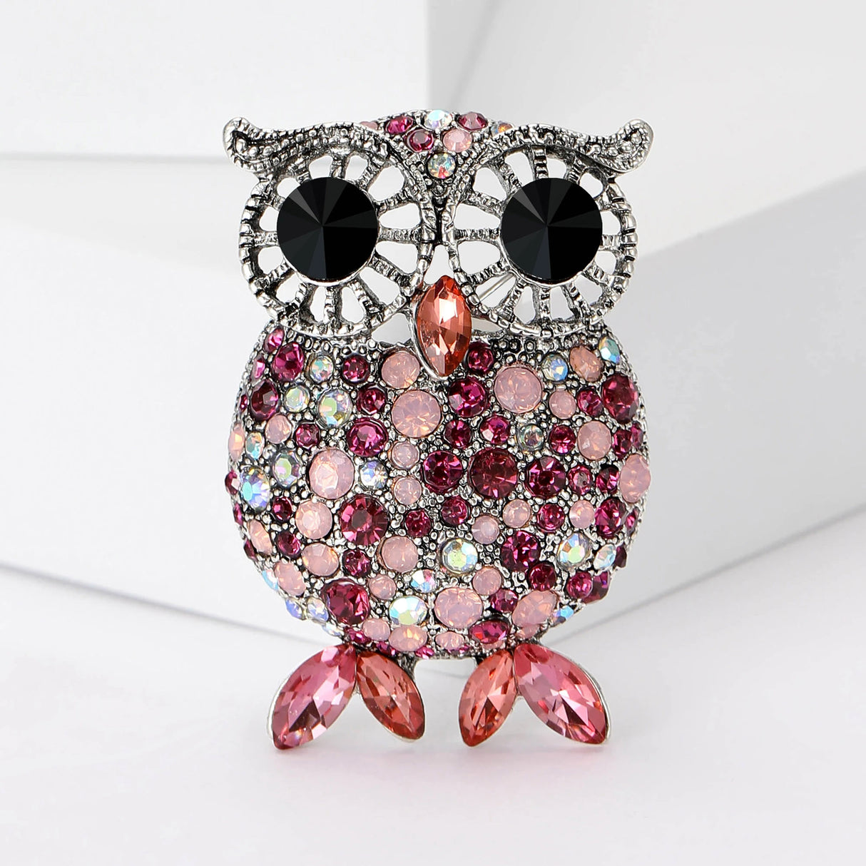 Owl brooch with silver-tone body, black eyes, and faceted stones in pink and iridescent tones, featuring pink pointed oval stones for feet, upright on white block.
