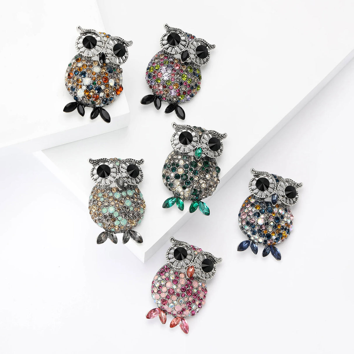 Set of six owl brooches in silver-tone metal with black eyes and multicolor faceted stones, each featuring varied stone colors and matching marquise-cut feet, flat lay on tiered white blocks.