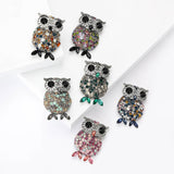 Set of six owl brooches in silver-tone metal with black eyes and multicolor faceted stones, each featuring varied stone colors and matching marquise-cut feet, flat lay on tiered white blocks.