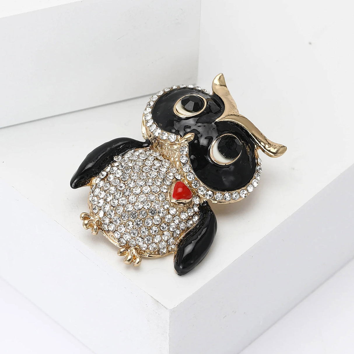 Owl brooch with black enamel wings and rhinestone eyes, clear pave stones on body, and red heart accent, all set in gold-tone metal. Angled top view on white studio backdrop.