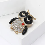 Owl brooch with black enamel wings and rhinestone eyes, clear pave stones on body, and red heart accent, all set in gold-tone metal. Angled top view on white studio backdrop.