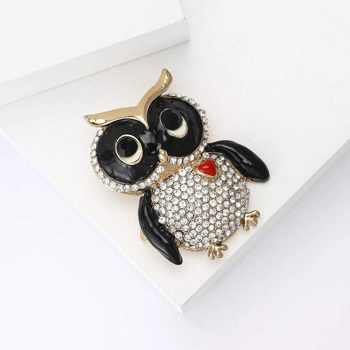 Owl brooch featuring black enamel details, clear faceted stones, gold-tone frame, and red heart-shaped accent, top-down view on white block.