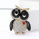 Owl brooch with black wings and eyes, body covered in small round clear stones, red heart accent and gold-tone frame. Standing pose on white background.