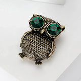 Owl brooch with textured gold-tone finish and large green faceted stone eyes, angled tow-down view on white studio background.