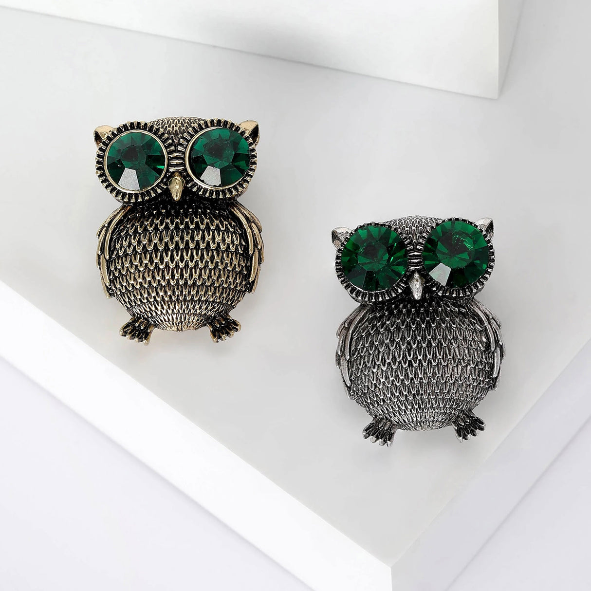 Set of two owl brooches with large green faceted eyes, one in gold-tone and one in silver-tone metal, top-down view on white background.