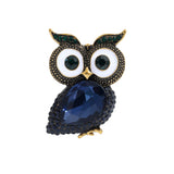 Owl brooch with dark blue faceted stone body, white eyes with dark green centers, and green accent stones on ears, cutout on white background.