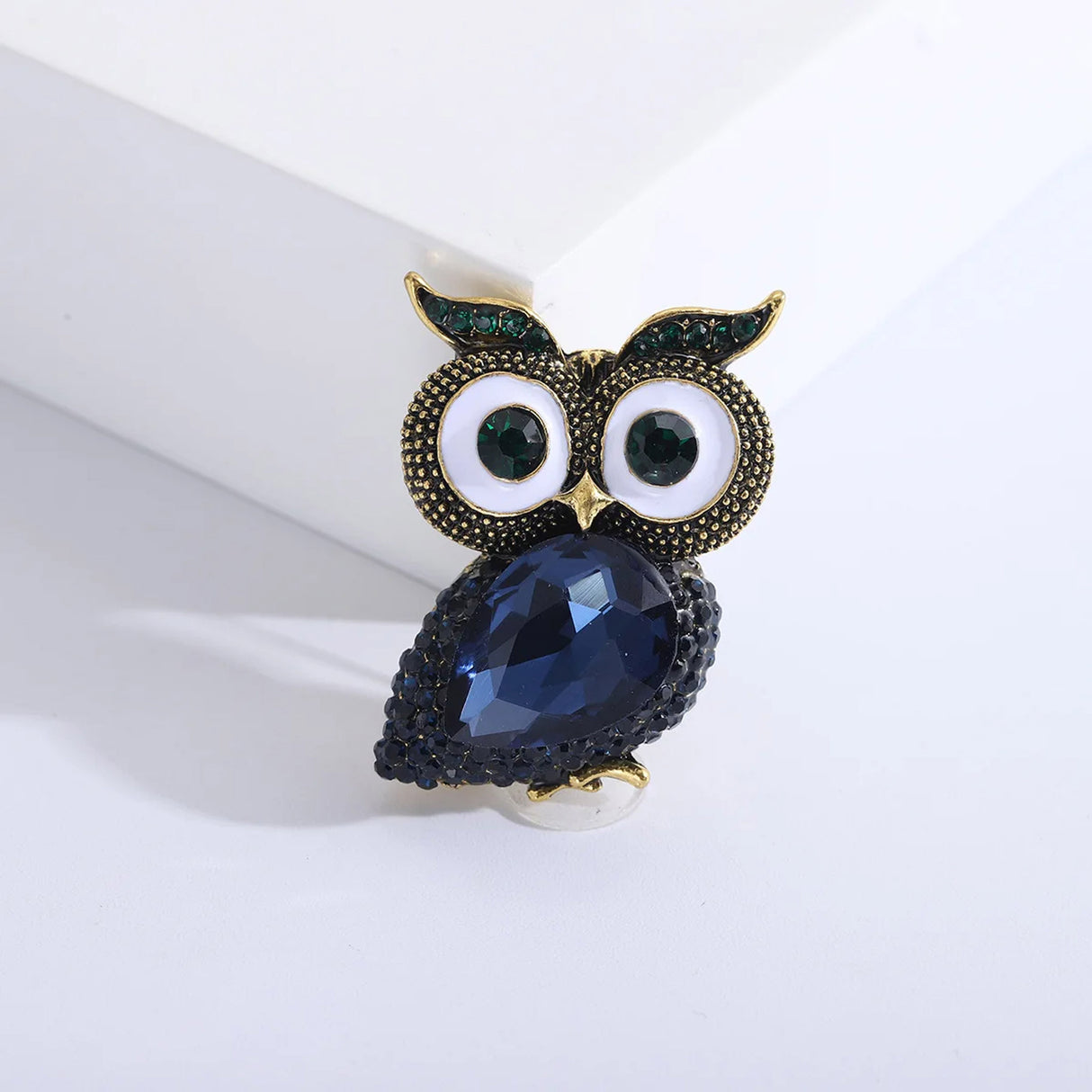 Owl brooch with dark blue faceted stone body, vintage-gold tone textured metal surface, and green accent eyes, upright on white background.